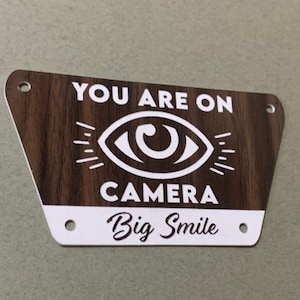 May include: A brown wooden sign with white text that reads "You are on camera Big Smile". The sign has a white eye graphic with eyelashes and a white circle in the centre.