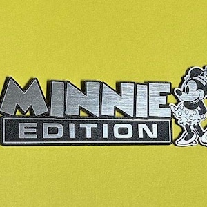 May include: A silver and black "MINNIE EDITION" emblem with a cartoon Minnie Mouse figure on a yellow background. The text is in a bold, blocky font, and the Minnie Mouse figure is in a classic pose.