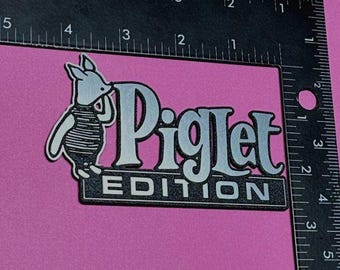 Może przedstawiać: Czarno-srebrne logo Piglet Edition z kreskówkowym prosiaczkiem w pasiastą koszulę. Prosiaczek patrzy w prawo i trzyma w pysku mały przedmiot.