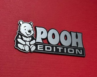 Może przedstawiać: Srebrny metalowy emblemat z czarno-białym rysunkiem animowanym Winnie the Pooh i tekstem "POOH EDITION".