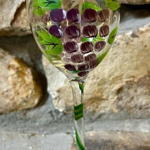 Op de afbeelding: Een helder wijnglas met een handgeschilderd druivenontwerp. Het glas heeft een tros paarse druiven en groene bladeren. De steel is versierd met groene strepen en de voet heeft een groen bladerenpatroon.