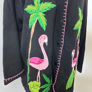 May include: A black knit cardigan featuring embroidered and sequined pink flamingos and green palm trees. The garment has a button-down front with black buttons and pink stitched detailing along the edges, cuffs, and placket.