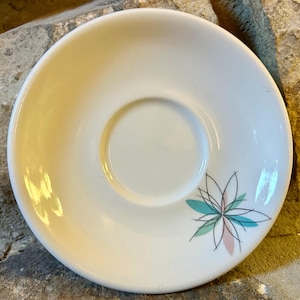 May include: White ceramic saucer with a central recessed area and a floral design. The flower has teal, pink, and black accents. The saucer is round and has a smooth surface.