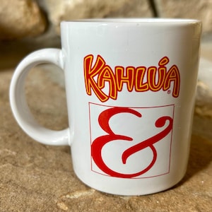 May include: White ceramic coffee mug with the Kahlúa logo in red and yellow. The logo includes the word "Kahlúa" and a red ampersand symbol. The mug has a curved handle and is set against a neutral background.