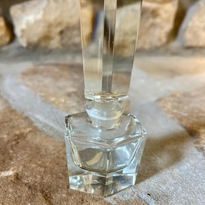 Puede incluir: Una botella de vidrio transparente con un tapón facetado. La botella está vacía y tiene forma cuadrada.