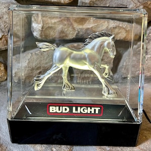 May include: A silver-colored horse figurine inside a clear acrylic display case. The base of the case is black, with the Bud Light logo in red and white. The horse is in a running pose.