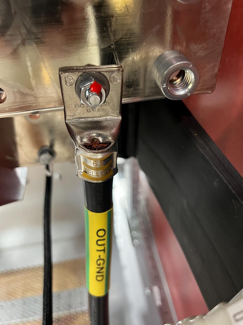May include: A black electrical wire with a yellow and green striped label that reads "OUT-GND". The wire is connected to a silver terminal block with a red button.