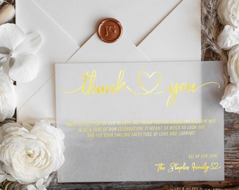 Op de afbeelding: Een witte envelop met een gouden zegel van was en een bedankkaart met gouden letters. De kaart zegt "Bedankt" met een hartje en een bericht waarin de gasten bedankt worden voor hun deelname aan de viering. De kaart is ondertekend met "The Staples Family".