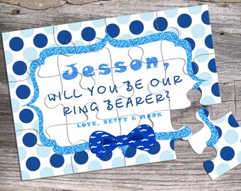 Op de afbeelding: Blauw en wit puzzel met een blauwe glitterrand. De puzzel zegt "Jesso, wil je onze ringdrager zijn? Liefs, Betty & Mark" met een blauwe vlinderdas met een snorpatroon.