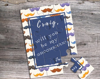 Op de afbeelding: Een legpuzzel met een marineblauw middenstuk met de tekst "Craig, will you be my groomsman?". De puzzelstukjes hebben een snorpatroon in verschillende kleuren. De puzzel ligt op een houten oppervlak.