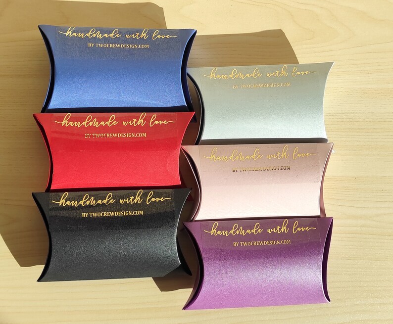 May include: Six pillow boxes in different colors, each with a gold sticker that says "handmade with love" and the website "BY TWOCREWDESIGN.COM".