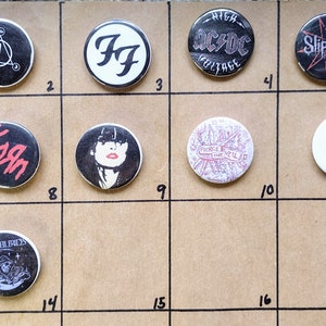 May include: A collection of 18 round button pins featuring various band logos, including Nirvana, Foo Fighters, AC/DC, Slipknot, The Cure, Korn, and more. The pins are displayed on a brown cardboard background.