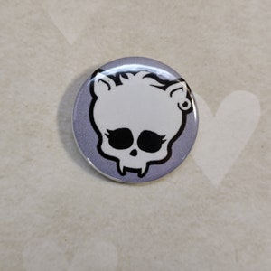 May include: A purple button with a white skull with black eyes and a black nose. The skull has a small black earring in its right ear. The skull has a small black mouth with two fangs.