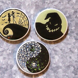 May include: Three round buttons with images from the Nightmare Before Christmas movie. The first button has a black silhouette of Jack Skellington and Sally on a hill with a spider web. The second button has a green background with a black silhouette of Oogie Boogie. The third button has a yin yang design with black and white silhouettes of Jack Skellington's head.