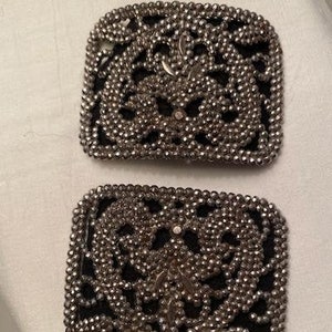 May include: Two silver-toned metal buckles with a black fabric backing. The buckles are square shaped and feature a decorative design of small beads and rhinestones.