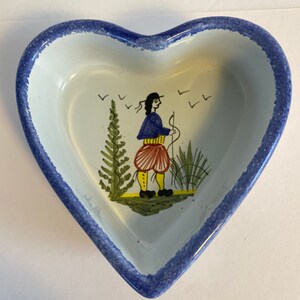 May include: A heart-shaped ceramic dish with a blue rim and a hand-painted design of a person in a blue jacket and red pants standing in a field with trees and birds.