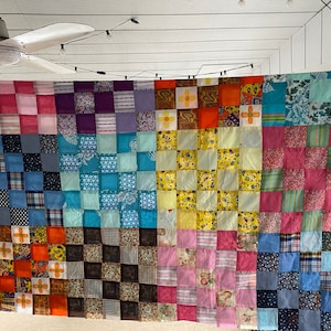 May include: A patchwork quilt made from a variety of colorful fabrics. The quilt is made up of squares of different colors and patterns, including pink, blue, green, yellow, and orange. The quilt is hanging on a wall.