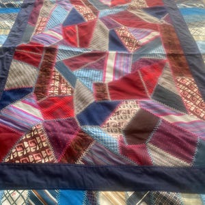 May include: A handmade quilt featuring a patchwork design of various fabrics in shades of red, blue, brown, and purple. The quilt has a dark blue border and is stitched with white thread. The quilt is made of repurposed neckties.