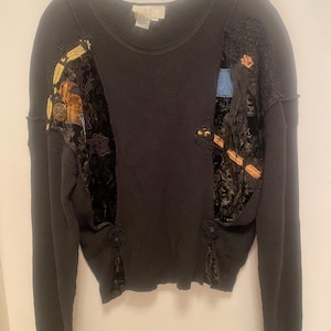 May include: Black long-sleeved waffle knit top with decorative patches of velvet, lace, and embroidered details. The patches feature gold, brown, and blue accents. The top has a round neckline and is hanging on a hanger.