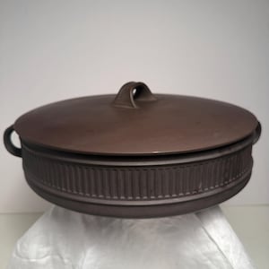 May include: A round, dark brown ceramic casserole dish with a matching lid. The exterior of the dish features a vertical ribbed texture around the side and two small handles on opposite sides. The lid has a central arched handle for lifting.