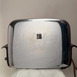 May include: A vintage chrome toaster with a rectangular shape and rounded corners. The toaster has a decorative etched design on the front and dark brown handles. A brown electrical cord extends from the base.