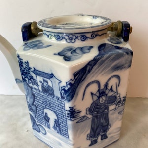 May include: A white porcelain teapot with blue and white painted designs. The teapot has a hexagonal body and a lid with two brass handles. The design features a Chinese landscape with figures and a building.