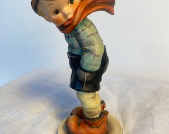 Puede incluir: Una figurita de porcelana de un niño con un gorro y una bufanda de color rojo y naranja, una camisa a cuadros azul y blanca y pantalones negros. El niño está de pie sobre una base blanca.