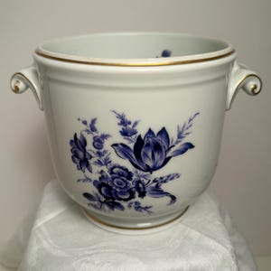 May include: A white porcelain cachepot featuring a hand-painted blue floral motif on the front. The vessel has two side handles with gold trim along the top rim and base. The interior is white, and the exterior displays a detailed botanical pattern.