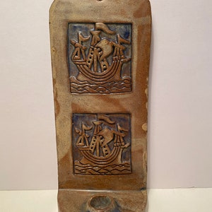 May include: A ceramic wall sconce with a candle holder and two raised images of ships with sails. The sconce is glazed in shades of brown, blue, and white.