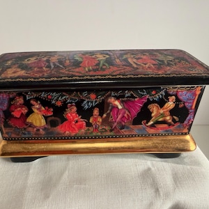 May include: A rectangular black box with a gold base and feet. The box features a detailed painting of figures in colorful costumes, with a gold trim. The top of the box has a painted scene of people dancing. The box is on a white surface.