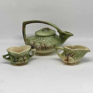 May include: A green and brown ceramic teapot and two matching creamer cups with a leaf and acorn design.