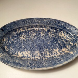 May include: An oval-shaped ceramic platter featuring a blue and white speckled pattern. The dish has a wide, slightly raised rim and a smooth, glossy finish, with the blue pigment creating a mottled, sponge-painted effect across the surface.