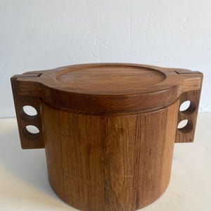 May include: A wooden ice bucket with a lid and two handles. The bucket is made of light brown wood with a natural grain pattern.