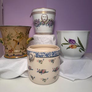 May include: A collection of four ceramic vessels including a beige planter with bird motifs, a white cylindrical pot with a relief face and floral trim, a white bowl with tulip illustrations, and a cream-colored jar with blue floral patterns.