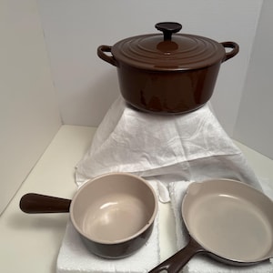 May include: A brown enameled cast iron Dutch oven with a lid, two small handles, and a black knob. Below it are a small saucepan and a frying pan, both with brown handles and cream-colored interiors. Kitchen cookware.