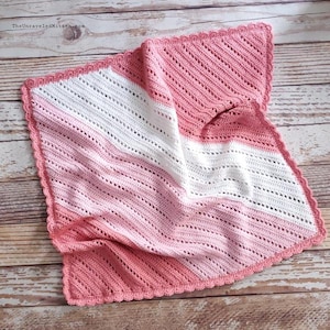May include: A handmade crochet baby blanket in shades of pink and white. The blanket features a diagonal striped pattern with a scalloped edge. The text "The Unraveled Mitten" is visible in the top left corner.