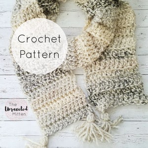 May include: A crochet scarf with a white and gray color gradient. The scarf has a fringe on each end and is made with a chunky yarn. The text "Crochet Pattern" is written on a white circle in the image. The text "The Unraveled Mitten" is written in a pink circle below the scarf.