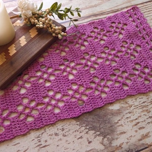 May include: A magenta-coloured, openwork crochet table runner. The runner has a repeating floral pattern and is displayed on a rustic wooden surface, highlighting its intricate design and texture. A wooden cutting board and floral accents are in the background.