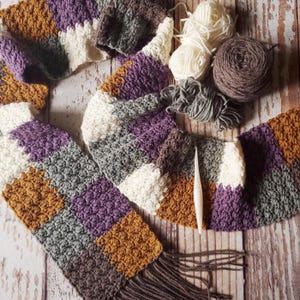 May include: A multi-coloured crocheted scarf with a fringe. The scarf is made with different shades of brown, purple, white, and grey yarn. The scarf is lying on a wooden surface with balls of yarn and a crochet hook.