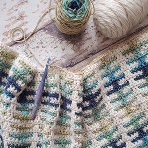Puede incluir: Una manta de ganchillo con un patr&oacute;n a rayas en tonos de azul, verde y blanco. La manta se est&aacute; trabajando con un gancho de ganchillo y lana.