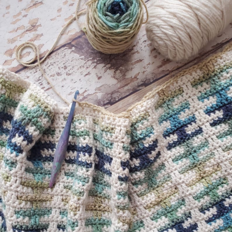 Puede incluir: Una manta de ganchillo con un patr&oacute;n a rayas en tonos de azul, verde y blanco. La manta se est&aacute; trabajando con un gancho de ganchillo y lana.