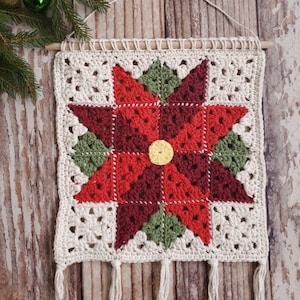 以下が含まれることがあります： 赤、緑、白のポインセチア柄のクロシェ編み壁掛け。上部に木製の棒、下部にタッセルが付いている。