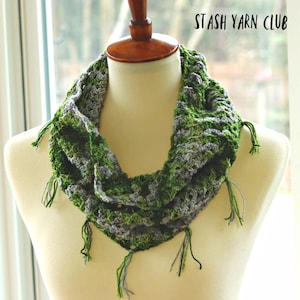 May include: A green and grey crocheted infinity scarf with fringe. The scarf is displayed on a white mannequin. The text "STASH YARN CLUB" is visible in the top right corner of the image.