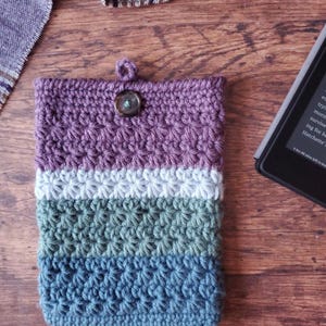 Pode incluir: Uma capa de telefone de crochê com um esquema de cores roxo, branco, verde e azul. A capa tem um fecho de botão e um design texturizado.