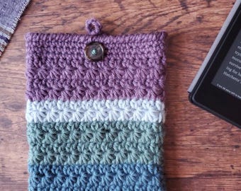 May include: A crocheted phone case with a purple, white, green, and blue color scheme. The case has a button closure and a textured design.
