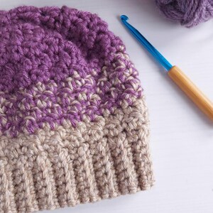 May include: A handmade crochet hat in shades of purple and beige. The hat is made with a textured stitch and has a ribbed brim. A blue crochet hook with a wooden handle and a ball of purple yarn are also in the image.
