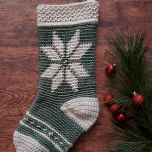May include: A crocheted Christmas stocking with a green and white snowflake design. The stocking is hanging on a wooden surface with a pine branch and red baubles.