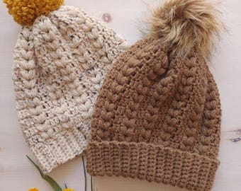 May include: Two handmade crochet beanies. One is beige with a mustard yellow pom-pom, the other is brown with a faux fur pom-pom. Both beanies have a textured stitch pattern and a ribbed brim. The beanies are displayed on a light-colored surface.