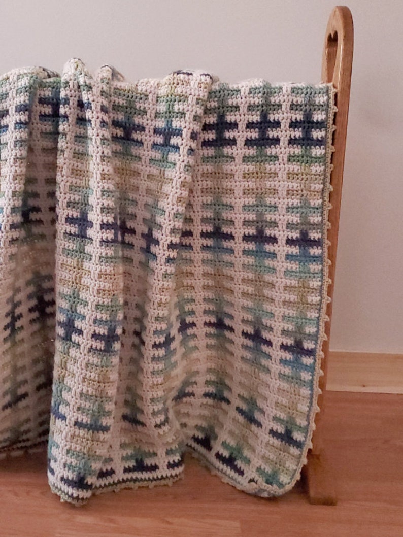 Puede incluir: Una manta de ganchillo con un patr&oacute;n geom&eacute;trico en tonos de azul, verde y blanco. La manta est&aacute; drapeada sobre una silla de madera.