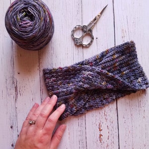 May include: A purple and grey knitted headband with a textured pattern. The headband is folded in half and is lying on a white wooden surface.